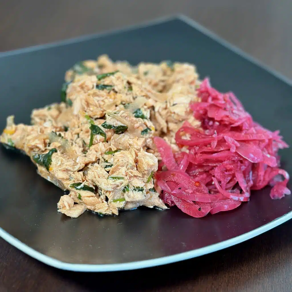 Black plater with a generous amount of Ginger Salmon Salad with a side of pickled onions, perfect for making tea sandwiches for brunch or adding to a lettuce salad